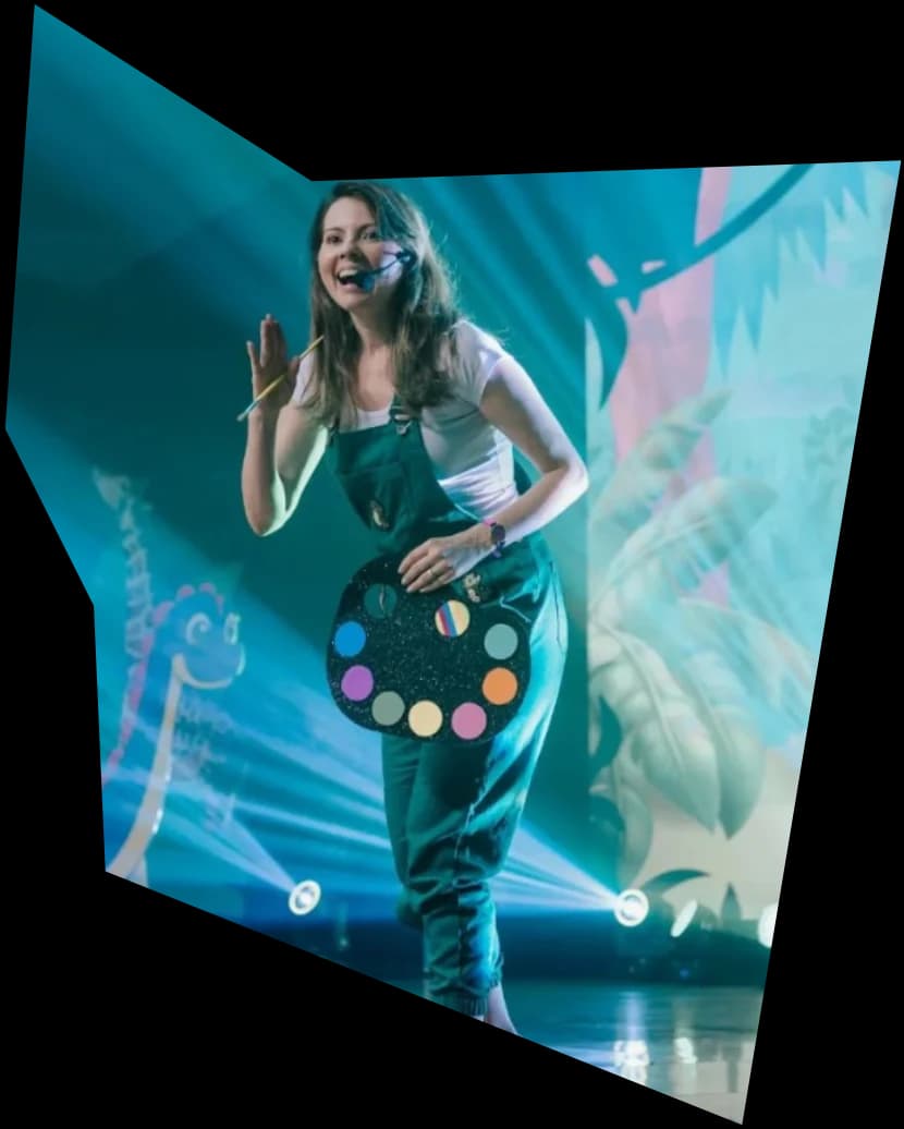Tia Pri no palco, sorrindo e acenando, com um macacão verde e uma paleta de tintas colorida